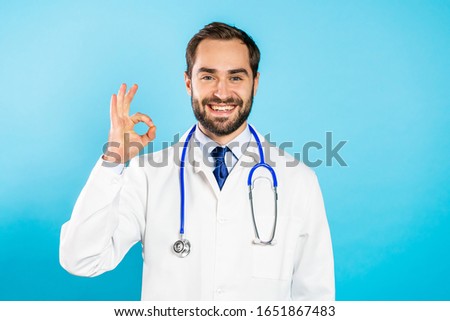 Portrait of smiling positive doctor in professional medical white coat showing OK sign gesture. Doc isolated on blue background.