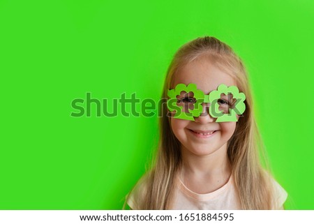 Cute Little blonde caucasian girl in mask of leprechaun shamrock clover glasses for irish St. Patrick's Day on green studio background. Mockup with copy space, template