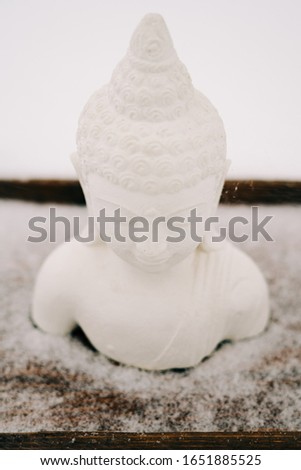 a small statue of a white Buddha made of plaster on a wooden stand during a snowfall in winter
