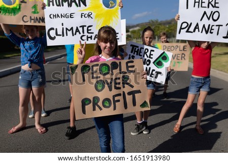 Front view of a group of Caucasian elementary school pupils walking down a road in the sun on a protest march, carrying signs with environmental and conservation slogans on them