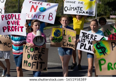 Front view of a diverse group of elementary school pupils on a protest march, carrying signs with environmental and conservation slogans on them, one girl shouting in a megaphone as they walk down a