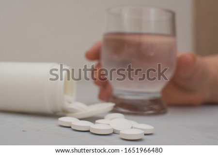 Woman holds a glass of water, pills are scattered on a gray table.