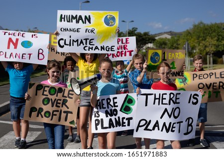 Front view of a diverse group of elementary school pupils walking down a road in the sun on a protest march, carrying signs with environmental and conservation slogans on them, one girl holding a
