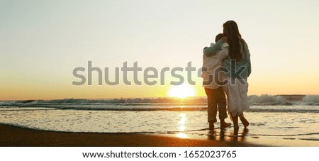 A woman hugging her son while standing on the beach surrounded by the sea during the sunset