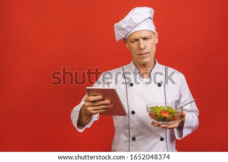 Picture of happy young senior chief cook in uniform standing isolated over red wall background, holding salad and tablet computer.