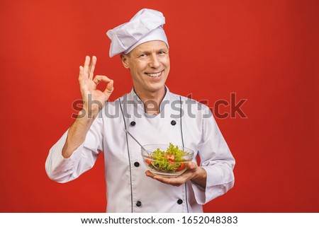 Picture of happy young senior chief cook in uniform standing isolated over red wall background, holding salad.