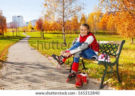 Happy blond 11 years old girl with amazing smile tie shoelaces on roller blades to go skating sitting on the bench in the autumn park on sunny day and turning face to camera