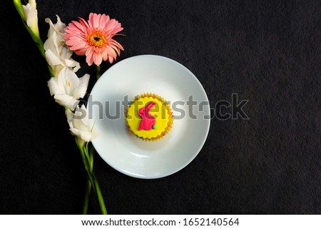 Pretty yellow vegan Easter cupcake with colorful pastel icing shape of Easter bunny rabbit on plate and spring Easter flowers on dark slate background, top view