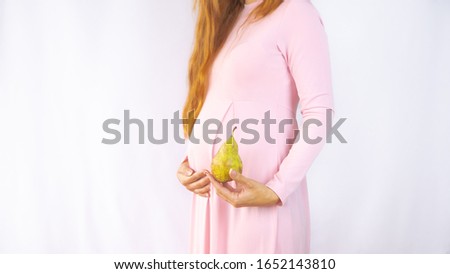 A close up photo of a pregnant young woman holding a pear next to her belly showing the size of the baby in a beautiful dress. Photos of fetal growth at 22 weeks pregnancy. Healthy pregnancy diet.