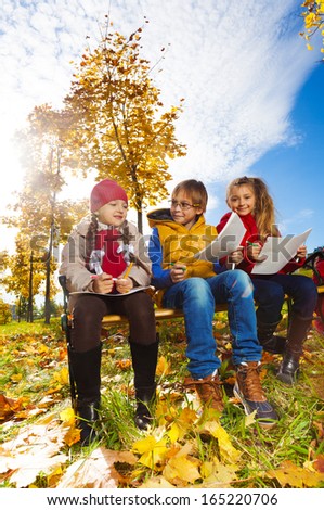 Group of three  kids, friends, boy and girls, sitting on the bench in autumn park on sunny day and draw pictures