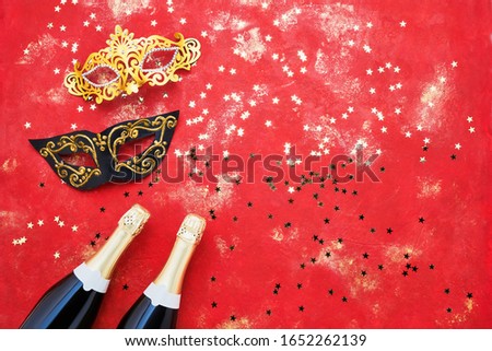 Two champagne bottles, carnival mask and golden confetti on red background. Flat lay of Purim Carnival celebration concept.