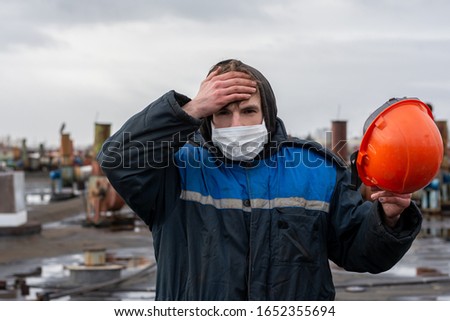 worker on the roof of the factory. holds a safety helmet in one hand
Feels unwell, headache. Holding his hand to his head. On the face is a white medical mask. Protection against bacteria, viruses.  Royalty-Free Stock Photo #1652355694