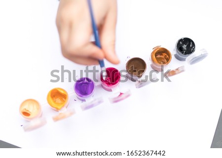 Pots of colorful paint in a paint set against white blank background with copy space pink purple orange brown a small child hand holding a paint brush dipping into paint pot macro close up
