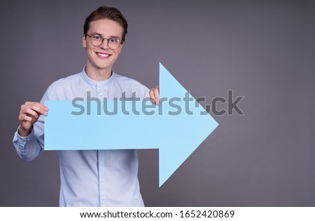 A serious charming young Caucasian guy, 20 years old, in a blue shirt, holding a large blue arrow in his hands, indicating the direction to the right, looking at the camera and smiling.
