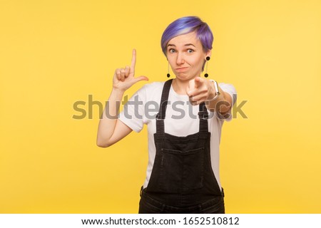You are loser! Portrait of hipster girl with violet short hair in denim overalls pointing to camera and showing loser lame gesture, making fun of your failure, unsuccess. yellow background studio shot