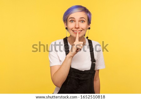 Shh, don't talk! Portrait of positive hipster girl with violet short hair in denim overalls showing silence gesture with fingers on lips, asking to be quiet. isolated on yellow background, studio shot