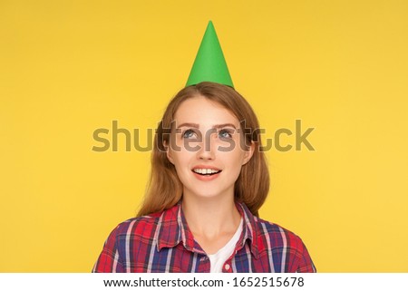 Closeup of happy dreamy ginger girl wearing funny party cone on head, looking up with dreamy expression and making wish on her birthday celebration. indoor studio shot isolated on yellow background
