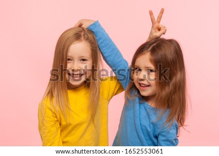 Happy carefree childhood, friendship. Portrait of two positive cute little girls showing bunny ears to each other, having fun, playing games together. indoor studio shot isolated on pink background