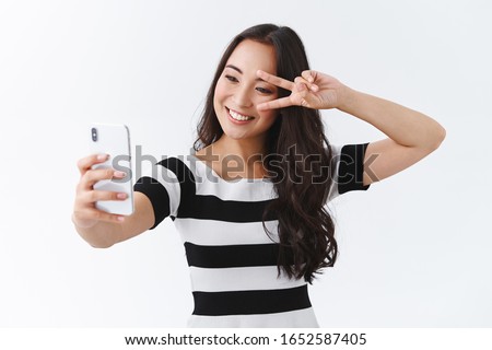 Cheerful, lively good-looking pretty asian woman in casual t-shirt, taking selfie on smartphone, show peace or victory sign near eye, making cute pose to photograph and send pic online