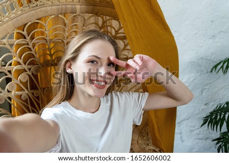 Happy and smiling young adult girl making self portrait, showing victory sign in camera and spending free time at home