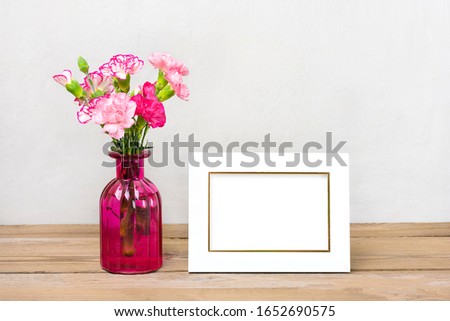 Small colored pink carnations in a vase and white photo frame,  on wooden background  and grey wall Hello spring, seasonal concept Mock up Place for text
