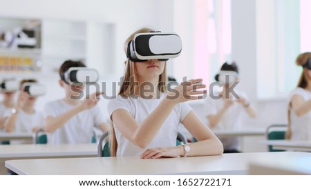 Portrait of a pretty girl sitting at first desk in the classroom. Future kids in white clothing wearing VR headsets for a game.