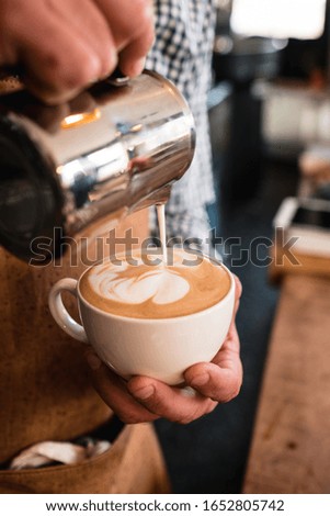 A vertical shot of a man pouring milk into a cappuccino cup in a cafe
