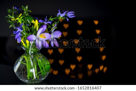 Blue yellow and green flowers bunch in a  transparent glass vase on a black table with many little hearts blurry bokeh in the background  