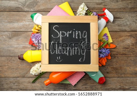 Blackboard with words Spring Cleaning, flowers and detergents on wooden table, flat lay