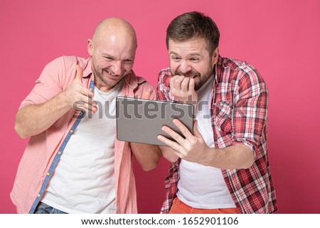 Two men laugh while looking at the tablet, they are shocked, see something funny on the screen and smile. Positive emotions.