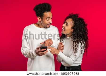 Young couple using mobile device. Man talking about apps and explains to girl how to use application. Red studio background