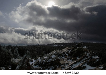 Photo of the "Wolfswarte" in the Harz Mountains. In the background is a snow-covered forest. The sky has a bright side on the left and a dark side on the right, with the sun in the middle.