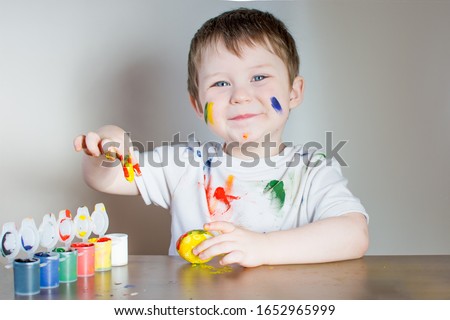 Happy blue-eyed child paints Easter eggs with paint. The boy on a light background in a white T-shirt, face, clothes and hands is painted with colorful paint. Royalty-Free Stock Photo #1652965999