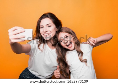 two sisters taking selfie in studio on orange background.
