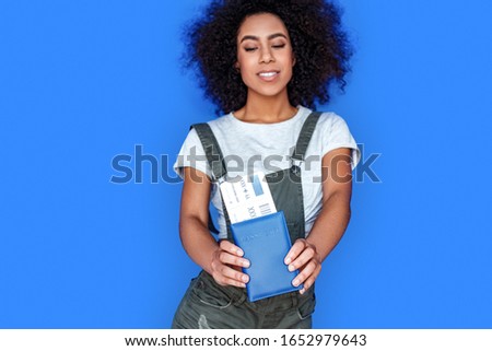 Young african american girl wearing denim overall standing isolated on blue wall looking down at passport and plane ticket close-up smiling happy blurred background