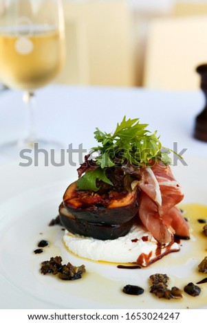 Food photography of grilled figs with proscuitot with rocket atop of ricotta cheese with a glass of white wine in the background set in a restaurant