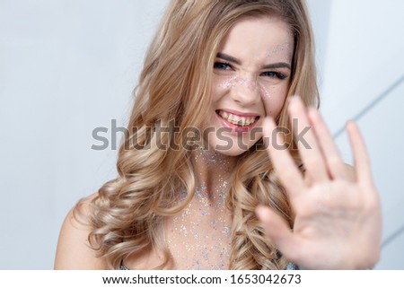 charming blonde with long curly hair makes a stop sign. Makeup with sequins on the face