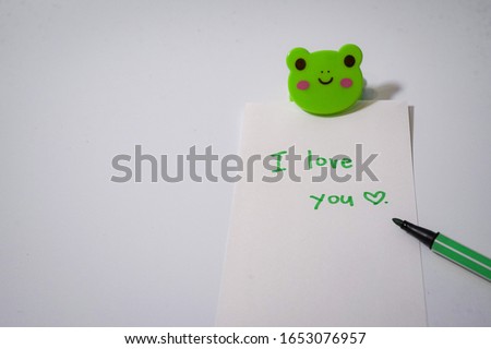 Someone wrote “ I love you “ in the piece of paper on the white plain walpaper with green pen. There is a cute frog clip on it.
