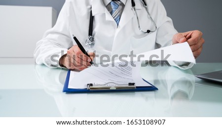 Doctor's Hand Signing Document On Desk In Clinic