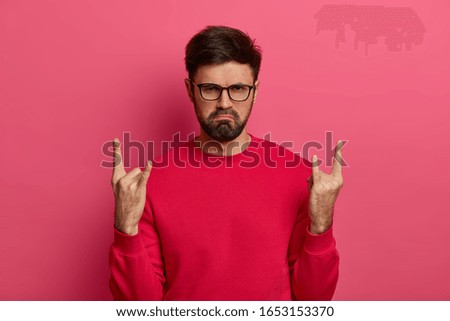 Rocking party. Gloomy dissatisfied unshaven man makes rock n roll gesture, enjoys great music, has charismatic face expression, feels cool, shows heavy metal sign, wears red sweater, optical glasses