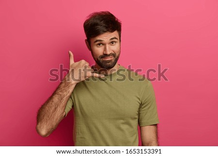 Handsome bearded man makes phone gesture near, asks girlfriend telephone number, says so call me maybe, has satisfied expression, dressed casually, poses against pink background. Body language