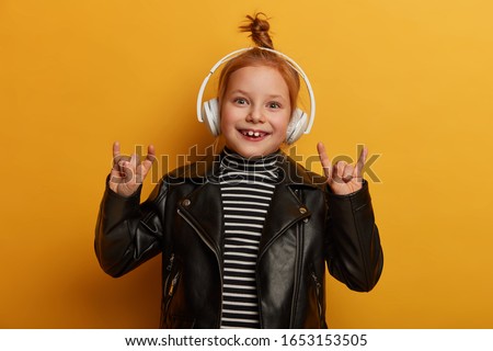 Optimistic kid rocker makes horn sign with fingers, enjoys listening heavy metal in headphones, has foxy hair knot, wears leather jacket, feels uplifted and overjoyed, chills and relaxes indoor