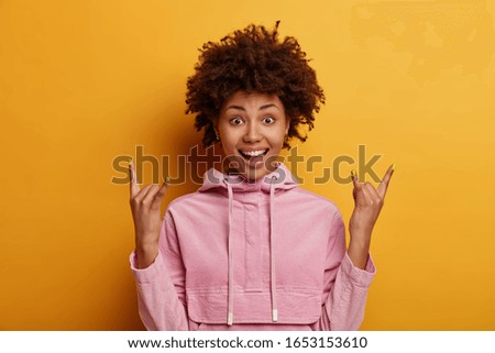 Horizontal shot of positive dark skinned woman makes rock sign, demonstrates rock n roll gesture, has good mood, wears casual sweatshirt, isolated on yellow studio wall. Body language concept