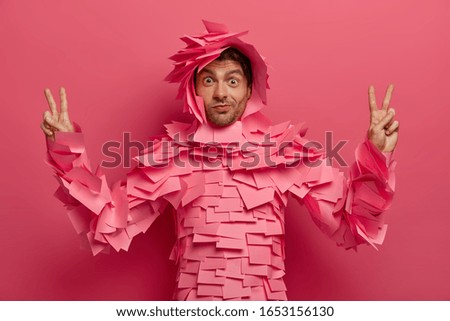 Surprised funny man has fun in office, poses in creative costume made of sticky notes, raises fingers in victory gesture, shows peace sign, isolated over pink background. Paper outfit. Monochrome