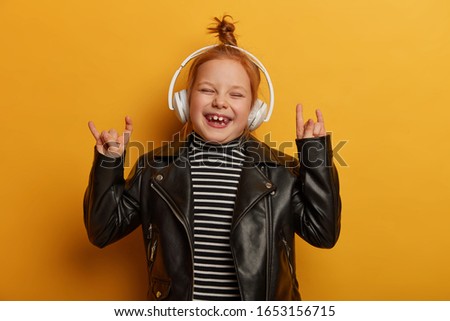 Happy small child rocker makes horn hand sign, rock n roll gesture, enjoys favourite music or melody in wireless headphones, wears leather jacket, giggles happily, isolated on yellow studio wall Royalty-Free Stock Photo #1653156715