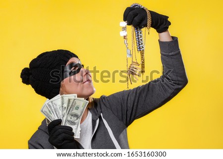 The concept of theft. A woman in a hat, mask and gloves holds a wad of dollars and jewelry in both hands, smiling at them. Yellow background and copy space