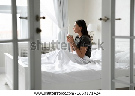 Smiling biracial young woman sit on white bed drink hot tea or coffee in the morning, happy African American female relax in bedroom enjoy warm beverage, good wake up in modern design home Royalty-Free Stock Photo #1653171424