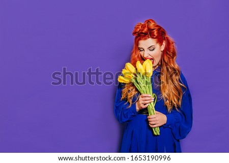 Beautiful girl in a blue dress with red hair on a purple background holds yellow tulips in her hands and does not look at the camera. Girl wants to eat tulips. Cool picture. Women's Day, 8 of march