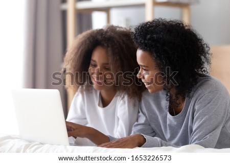 African american mom and teen daughter using laptop talk do online shopping chatting in social media lying on bed, smiling black mother and teenage girl order buy in internet on computer in bedroom