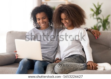 Happy african american family young mother and teen daughter using laptop computer sitting on couch, smiling mixed race parent mom with teenager girl having fun watching movie online at home together